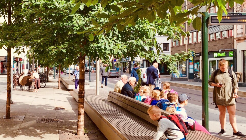 VENTER PÅ TRIKKEN: Her kan barn og eldre slappe av mens de venter på trikken. Her er det satt opp et hele 60 meter langt flerfunksjonsmøbel til å sitte, ligge, kjøre lekebil på eller det kan fungere som en catwalk på markedsdager. En liten gruppe barnehagebarn har tatt turen til byen sammen med førskolelæreren som sjekker klokka.Bymøbelet har en sokkel av varmgalvanisert og lakkert stål og trespiler av Keboney Radiata. (Foto: Bjørbekk & Lindheim)