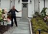 Mann på steingang foran et grått hus med blomster og grønne planter.